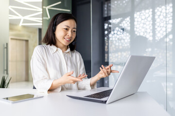 Successful smiling businesswoman working inside office at workplace, using laptop for video call, smiling. An Asian woman uses a headset to consult and communicate with a client.