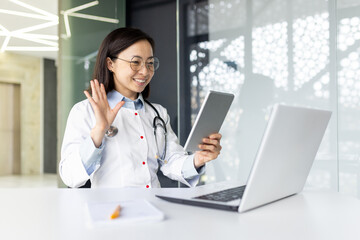 Young Asian female doctor working inside medical office consulting patients remotely, smiling with...