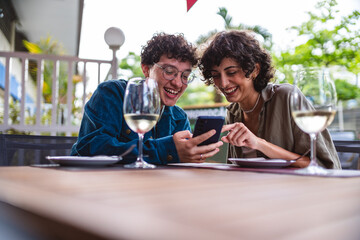 Jovem casal sentado a mesa de um restaurante ao ar livre conversando, usando smartphone.