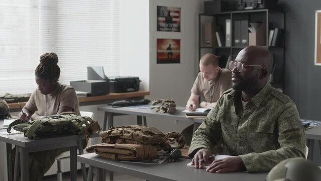 Side shot of cadet in glasses and camouflage uniform answering question and then writing during military service class in academy