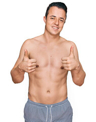 Fototapeta premium Handsome young man wearing swimwear shirtless success sign doing positive gesture with hand, thumbs up smiling and happy. cheerful expression and winner gesture.