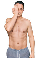 Fototapeta premium Handsome young man wearing swimwear shirtless covering one eye with hand, confident smile on face and surprise emotion.