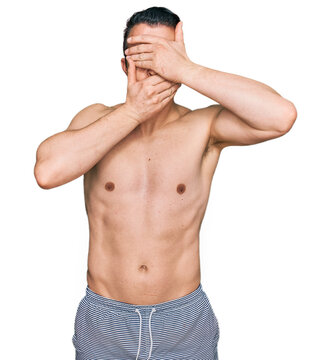 Handsome young man wearing swimwear shirtless covering eyes and mouth with hands, surprised and shocked. hiding emotion