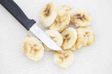 Bananas on white cutting board. Ripe banana slices isolated on white. Sunlight fruits. Quick healthy morning snack. Yellow fruits with brown stains. Cut banana slices. Small kitchen knife.	