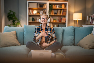 Obraz premium A mature woman listens to a guided meditation on headphones, she is on the couch in her apartment and relaxes