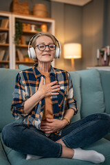 Obraz premium A mature woman listens to a guided meditation on headphones, she is on the couch in her apartment and relaxes