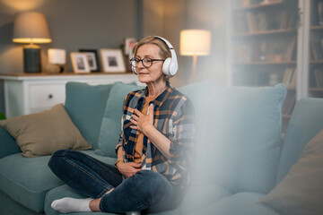 A mature woman listens to a guided meditation on headphones, she is on the couch in her apartment and relaxes