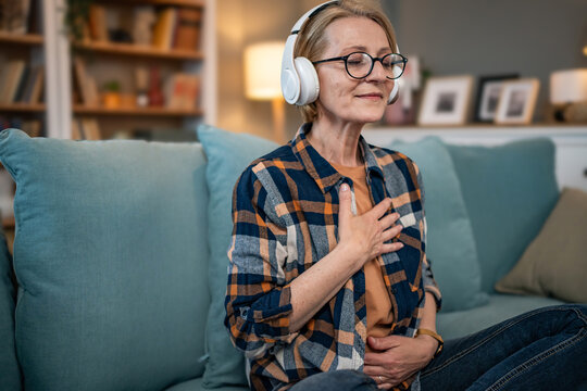 A mature woman listens to a guided meditation on headphones, she is on the couch in her apartment and relaxes