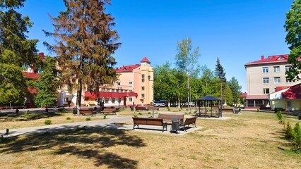 Around the city square with grass lawns, trees and wooden benches are buildings with metal tile...
