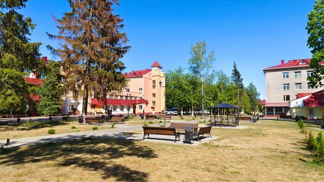 Around the city square with grass lawns, trees and wooden benches are buildings with metal tile roofs and poles with lanterns. In the middle of the public garden there is a gazebo Summer sunny weather