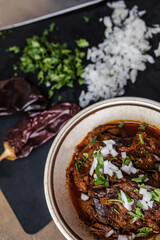 A bowl of slow cooked and braised birria beef meat in a vintage bowl with chopped onions, cilantro, guajillo peppers, and broth