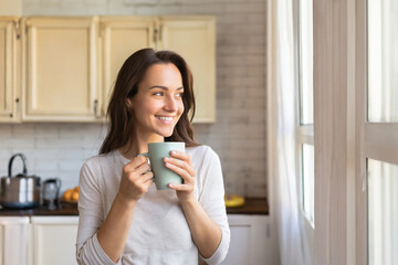  young adult woman at home in the kitchen with a cup in hand, coffee or tea, smiling happy relaxed