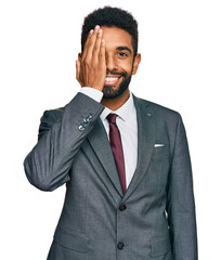 Young african american man wearing business clothes covering one eye with hand, confident smile on face and surprise emotion.