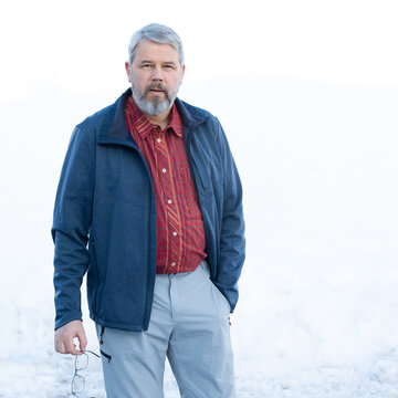 Man, 56 years old with gray hair and a gray beard, standing in front of a snowy area in winter, wearing a red checked shirt, dark blue jacket, light gray trousers, glasses in his hand