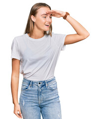 Fototapeta premium Beautiful young blonde woman wearing casual white t shirt very happy and smiling looking far away with hand over head. searching concept.