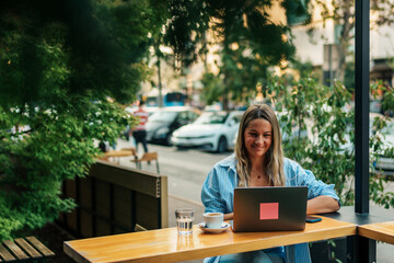 Seated in the café's outdoor area, a woman in her thirties, a freelancer, diligently works on her laptop, surrounded by the garden's tranquility.