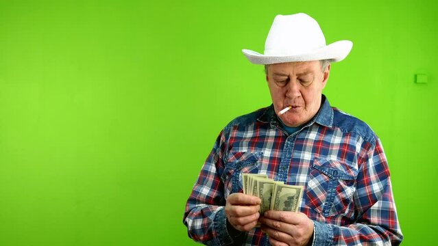Senior man in white cowboy hat sorts dollars, smokes cigarette and blows smoke.