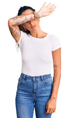 Young hispanic woman with tattoo wearing casual white tshirt covering eyes with arm, looking...
