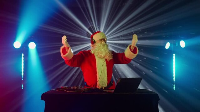 DJ Santa Claus mixing tracks in a nightclub at a Christmas and New Year party or Corporate events. Senior disc jockey as Santa listening music, headphones, laptop, mixer controller player, turntable.