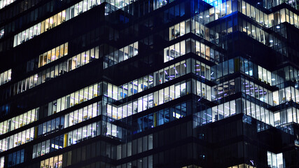 Obraz premium Fragment of the glass facade of a modern corporate building at night. Modern glass office in city. Big glowing windows in modern office buildings at night, in rows of windows light shines. 