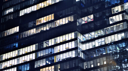 Obraz premium Fragment of the glass facade of a modern corporate building at night. Modern glass office in city. Big glowing windows in modern office buildings at night, in rows of windows light shines. 