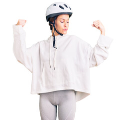 Beautiful brunette young woman wearing bike helmet and sporty clothes showing arms muscles smiling proud. fitness concept.
