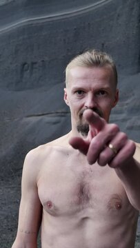 Vertical screen: portrait of a young handsome guy with a naked body pointing his finger at the camera and looking menacingly. A menacing angry man points his finger forward. Scary portrait, danger