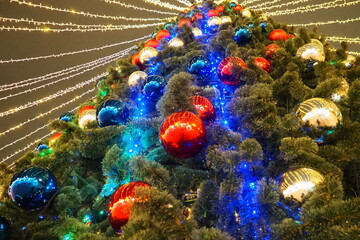 Multi-colored glass shiny balls on the New Year's city tree. Glowing garlands hang over the...