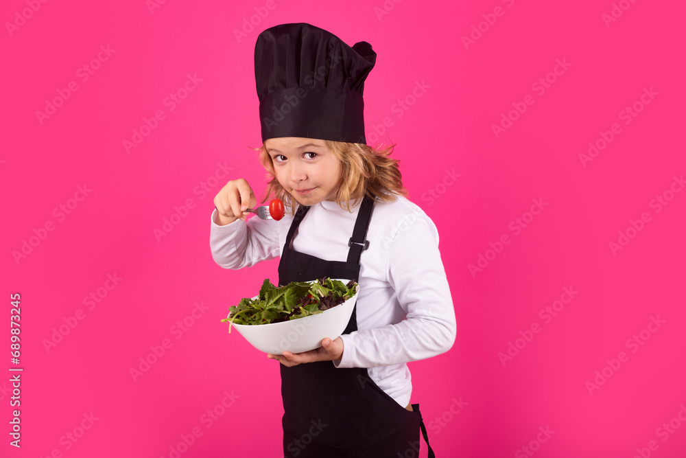 Wall mural Kid cook hold plate with vegetables and fork with tomato. Child chef cook prepares food on isolated studio background. Kids cooking. Teen boy with apron and chef hat preparing a healthy meal.