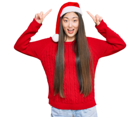 Young chinese woman wearing christmas hat smiling amazed and surprised and pointing up with fingers and raised arms.