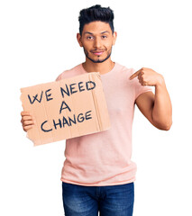Handsome latin american young man holding we need a change banner pointing finger to one self...