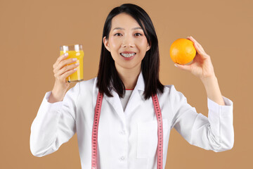 Female Asian nutritionist with orange and glass of juice on beige background