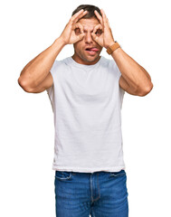 Naklejka premium Handsome muscle man wearing casual white tshirt doing ok gesture like binoculars sticking tongue out, eyes looking through fingers. crazy expression.