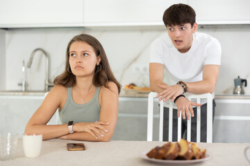 Sad young girl sitting at the table while her boyfriend is quarreling with her standing behind in...