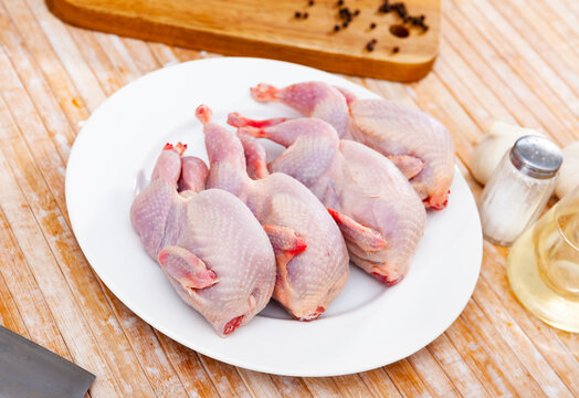 Uncooked quails on white plate on wooden table. Home kitchen, preparing food.