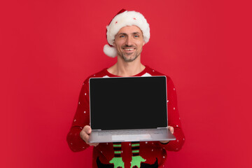 Middle aged santa hold laptop with blank screen. Santa with laptop. Man in Christmas winter sweater on red background.