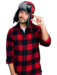 Hispanic young man with beard wearing fluffy earmuff hat smiling and confident gesturing with hand doing small size sign with fingers looking and the camera. measure concept.