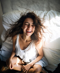 Happy young single brunette woman smiling and laughing alone in her bed in early morning, sunlight falling on her face from window, high angle