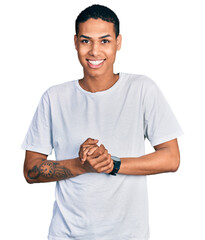 Young hispanic man wearing casual white t shirt with hands together and crossed fingers smiling relaxed and cheerful. success and optimistic