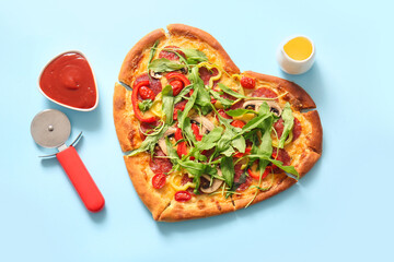 Tasty heart shaped pizza with bowl of ketchup and cutter on blue background