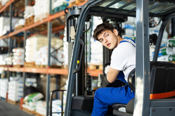 Fototapeta na wymiar Guy works on fork lift loader in construction rack area warehouse store repair materials. Young male employee looks back, chooses safe route for movement of equipment mechanism