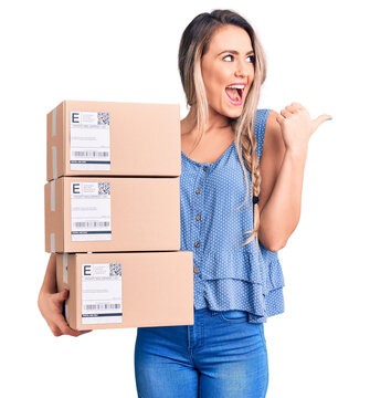 Young beautiful blonde woman holding delivery boxes pointing thumb up to the side smiling happy with open mouth