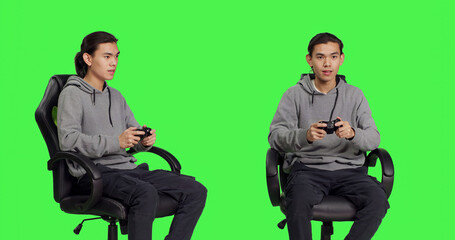 Asian guy playing videogames over greenscreen backdrop, using controller and having fun with...