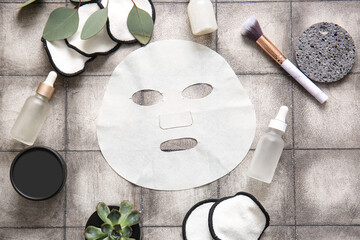 Facial sheet mask with different cosmetic products and houseplant on grey tile background