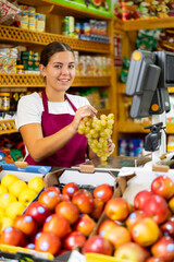 Obraz premium Positive young woman holding bunch of ripe grapes offering fresh fruits and looking in camera at greengrocery store