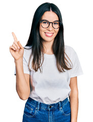 Young hispanic woman wearing casual clothes and glasses showing and pointing up with finger number one while smiling confident and happy.