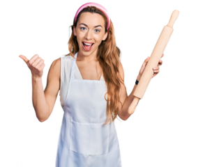 Young caucasian woman wearing professional baker apron holding kneading roll pointing thumb up to the side smiling happy with open mouth