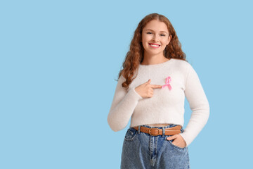 Young redhead woman pointing at pink ribbon on blue background. Breast cancer awareness concept