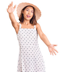 Young beautiful chinese girl wearing summer hat looking at the camera smiling with open arms for hug. cheerful expression embracing happiness. - obrazy, fototapety, plakaty