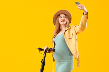 Young pregnant woman with electric kick scooter taking selfie on yellow background - obrazy, fototapety, plakaty
